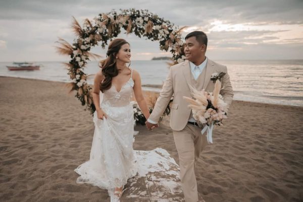 A married couple looking at each other while walking towards the camera. The bride is holding onto her dress and the groom is holding a bouquet of flower. It is a beach wedding with a circle arch decorated with flowers and leaves.