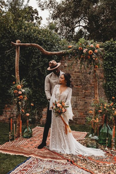 A married couple in a bohemian style wedding theme. The bride and groom are dressed in bohemian style posing for a photo with the rustic backdrop at the background. 