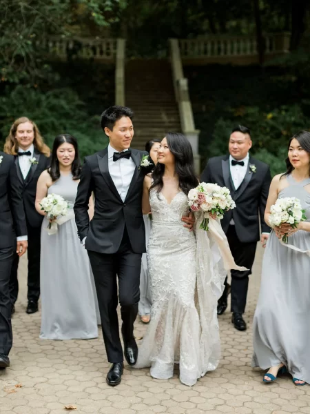 A married couple is walking towards the camera while looking at each other. The bride is holding a bouquet of flower. All the groomsmen and bridesmaid were standing at the back and looking at the couple.