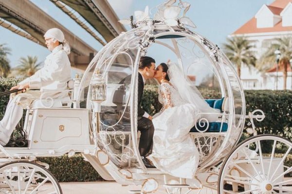 A fairytale wedding theme. A married couple is kissing inside the transparent Cinderella themed carriage while a driver is driving the carriage. The driver is dressed up in 1800s fashion.