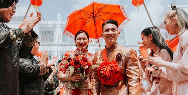 A married couple (Jane Chuck and Han Pin Ma) is dressed in traditional chinese wedding attire with an umbrella behind them smiling and walking towards the camera. The groomsmen and bridesmaid were dressed in traditional Chinese attire as well standing at the side looking at the couple. 