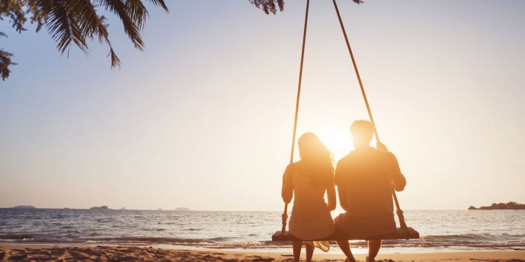 A couple is enjoying their honeymoon destination at the beach. They are seating on a swing with their back facing the camera; looking at the view of the ocean waiting for the sunset.